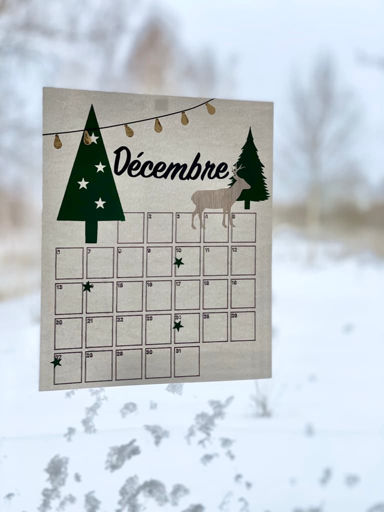 Calendrier de décembre décoré de sapins verts et rouges suspendu devant une fenêtre enneigée