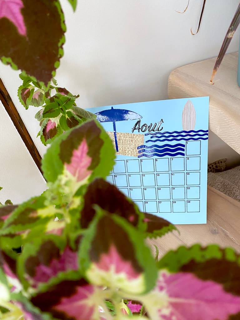 Calendrier du mois d’août décoré en bleu et placé derrière une plante aux feuilles roses et vertes