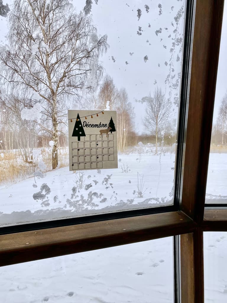 Calendrier de décembre accroché à une baie vitrée devant un paysage de forêt enneigée