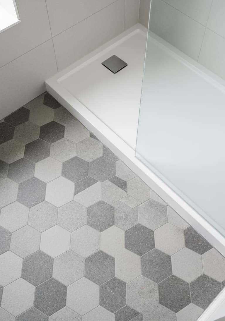 Vue plongeante sur un sol de salle de bain rénové avec des carreaux hexagonaux gris et le coin d'un receveur de douche neuf.