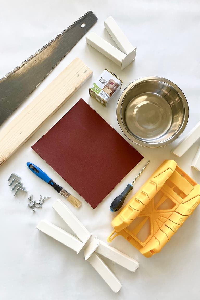 Scie, tasseaux de pin, papier abrasif, équerres, vis, gamelle en inox et boîte à onglets posés sur une table blanche