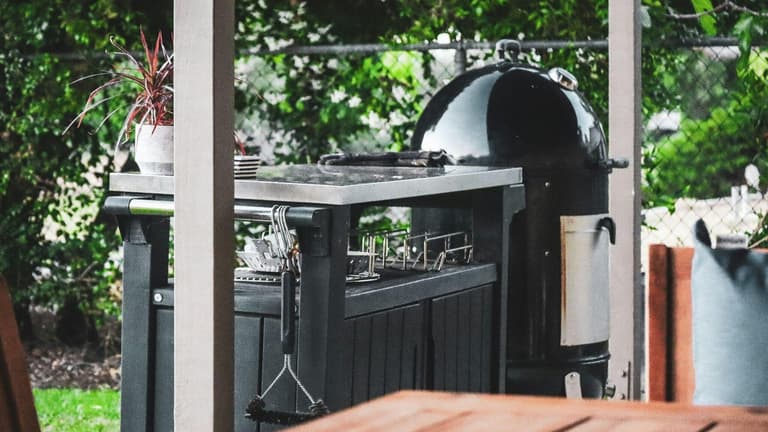 Cuisine extérieure noire avec plan de travail en inox et barbecue sous une terrasse couverte donnant sur un jardin.