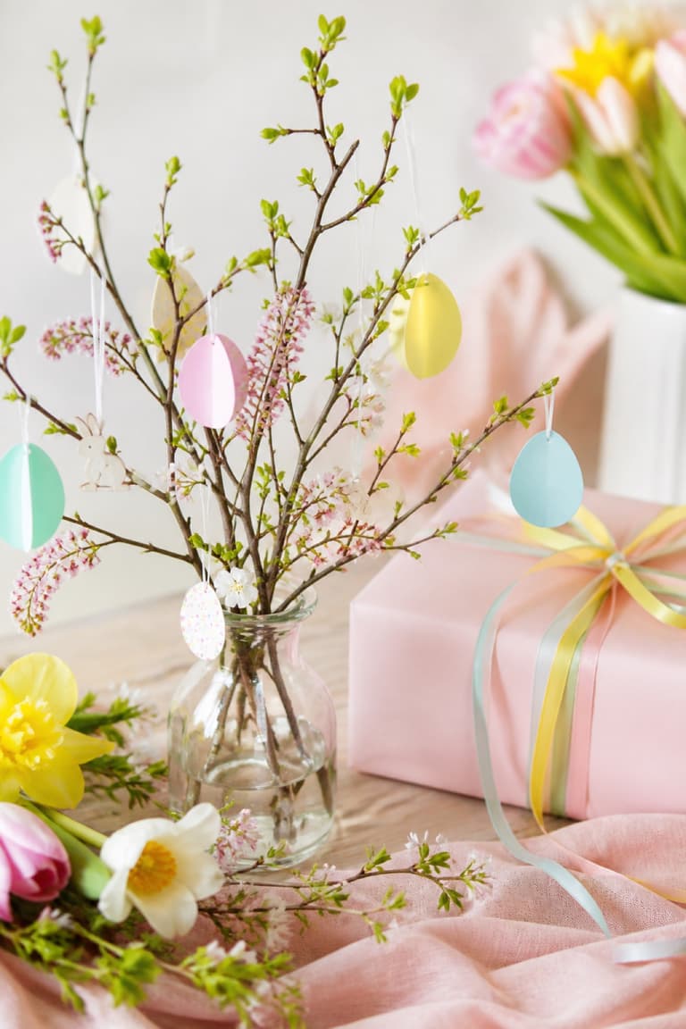 Branche fleurie décorée d’œufs pastel suspendus et d’un cadeau emballé dans des rubans colorés