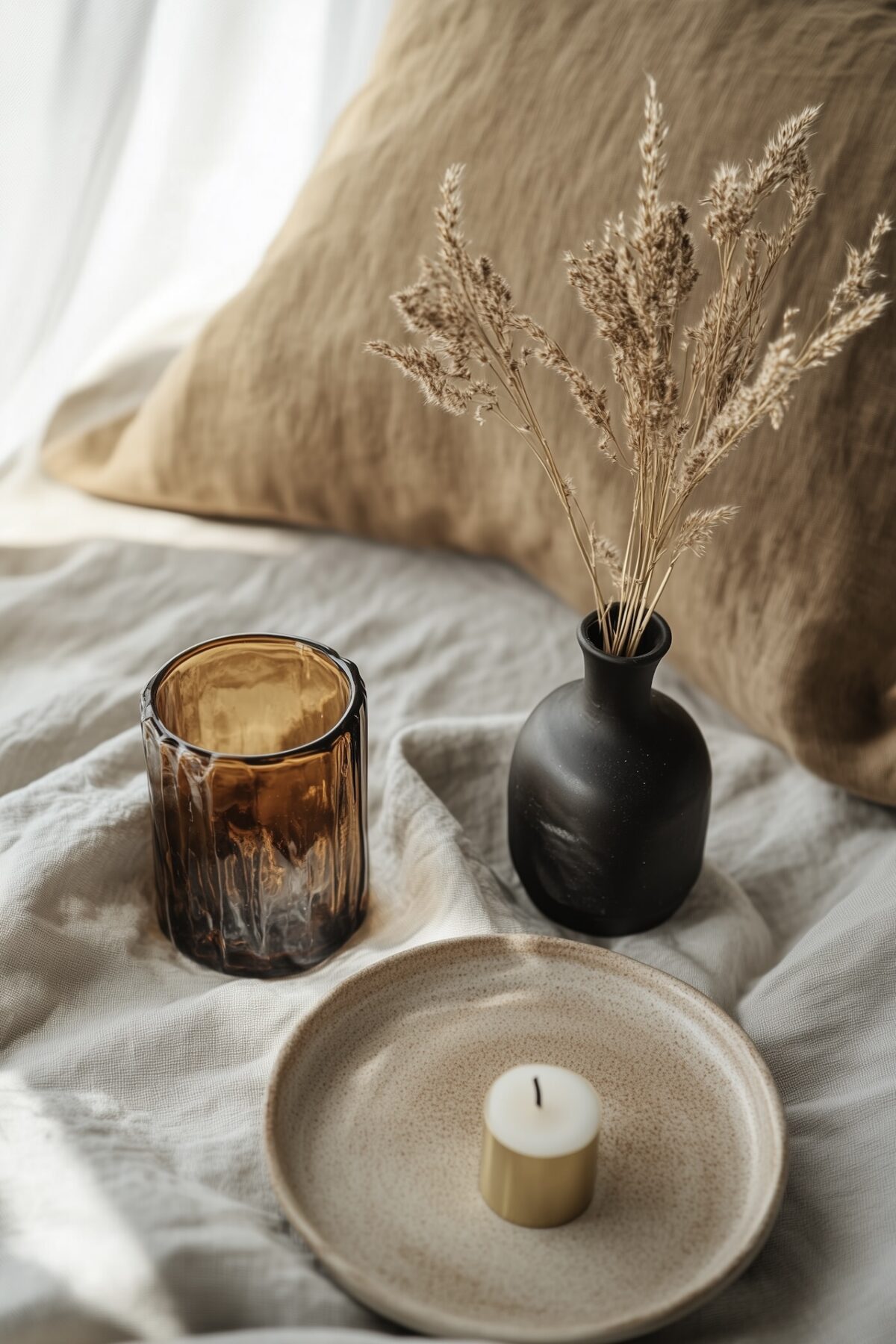 Vignette décorative sur canapé en lin avec coussin camel, verre texturé ambré, petit vase noir mat avec graminées séchées, coupelle en grès artisanal avec bougie, lumière douce naturelle
