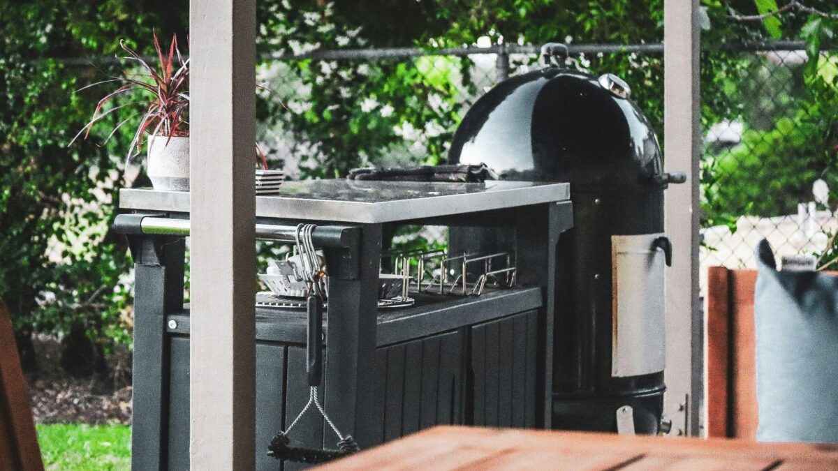 Cuisine extérieure noire avec plan de travail en inox et barbecue sous une terrasse couverte donnant sur un jardin.